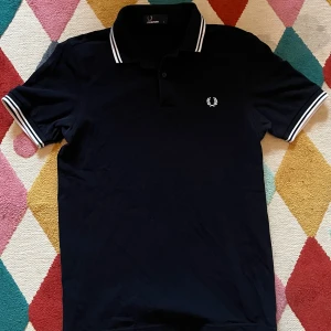 Svart Fred Perry pikétröja S - Svart pikétröja från Fred Perry i storlek S. Klassisk design med vit broderad logga på bröstet och vita ränder på krage och ärmslut. Tröjan har korta ärmar och två knappar vid halsen. Perfekt för dig som gillar stilrena och sportiga plagg. Använd 1-3 gånger 