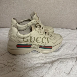 Gucci Rhyton sneakers i beige skinn - Chunky Gucci Rhyton sneakers i beige skinn med klassisk logga och gröna/röda ränder på sidan. Skorna har rund tå, snörning och en rejäl platt sula som ger en cool streetstyle-vibe. Perfekta för dig som vill sticka ut med lyxig känsla och trendig design.