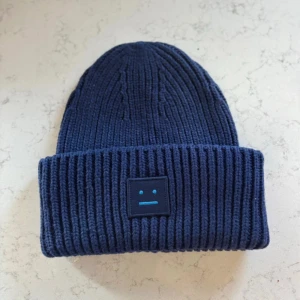 Mörkblå ribbstickad mössa Acne Studios - Säljer en mörkblå ribbstickad mössa från Acne Studios med det ikoniska Face-logotypen framtill. Mössan är i tjockt, mjukt material och har bred uppvikt kant. Perfekt för kalla dagar och ger en clean, stilren vibe till din outfit.