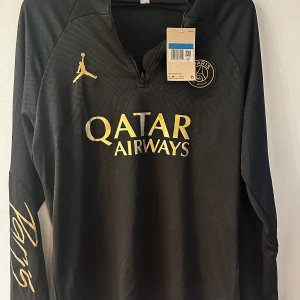 PSG tracksuit ny! - Snygg svart långärmad PSG fotbollströja från Jordan med guldfärgade detaljer och loggor. Tröjan har dragkedja vid halsen, Paris Saint-Germain-emblem på bröstet och Qatar Airways-tryck i guld. Tillverkad i lätt och funktionellt material och byxorna medföljer