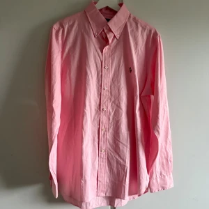 Rosa skjorta från Ralph Lauren - Klassisk rosa skjorta från Ralph Lauren med button down-krage och broderad logga på bröstet. Skjortan är långärmad, har normal passform och är tillverkad i mjuk bomull. Perfekt för dig som gillar stilrena och tidlösa plagg.