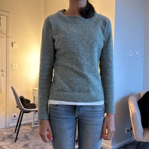 Grön/grå stickad tröja med rund hals - Mysig grå/grön stickad tröja med rundad hals och ribbade muddar vid ärmslut och nederkant. Tröjan har en enkel, klassisk design och är långärmad, perfekt för kyliga dagar. Passformen är normal och materialet känns mjukt mot huden.