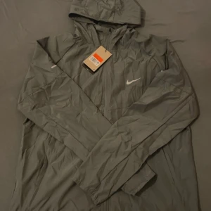 Grå vindjacka från Nike med huva - Grå vindjacka från Nike med vit logga på bröstet och huva.  