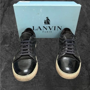 Svarta skor från Lanvin - Snygga svarta sneakers från Lanvin med glansig tå i skinn och ovandel i mocka. Skorna har svarta snören, rund tå och vit platt sula med diskret logga undertill. Perfekta för dig som gillar stilrena och exklusiva sneakers med en lyxig touch.