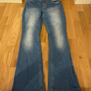 Blå bootcut jeans från Street One - Snygga blå jeans från Street One med bootcut passform och hög midja. Jeansen har knäppning med fyra synliga knappar och klassiska fickor. Materialet är denim med en lätt tvättad look och ljusare partier framtill. Perfekta för dig som gillar retrovibbar och bekväm stil.