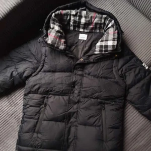 Svart Burberry pufferjacka med huva - Säljer en svart pufferjacka från Burberry med stor patch på ärmen där det står 'BURBERRY LONDON ENGLAND'. Jackan har huva med gråa och vita ränder på insidan, dragkedja och knappar framtill. Perfekt för kalla dagar och riktigt snygg.