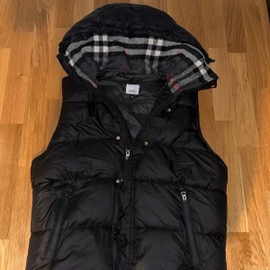 Svart dunväst från Burberry med huva - Svart jacka/väst från Burberry med avtagbar huva och klassiskt rutigt foder i grått, vitt och rött. Västen har dragkedja, tryckknappar och två fickor med dragkedja framtill. Stor Burberry-patch på ena axeln. Perfekt för kallt väder plus är väldigt stilig.
