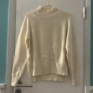 Krämvit stickad polotröja med slits - Mysig krämvit stickad polotröja med ribbad hög krage och breda ribbade muddar. Tröjan har lång ärm och snygg slits på båda sidor  för extra detalj. Perfekt för lager-på-lager och chill stil under kyliga dagar. Endast testad