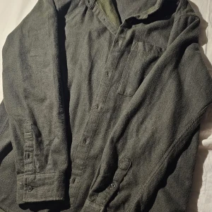 Mörkgrön overshirt i ullmix - Säljer en mörkgrön overshirt med klassisk krage och bröstficka. Skjortan har knäppning framtill och långa ärmar med manschett. Materialet är en mjuk ullmix som ger en skön och avslappnad känsla. Perfekt att bära lager på lager under kyligare dagar.