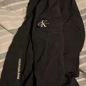 Svart vindjacka från Calvin Klein Jeans - Svart vindjacka från Calvin Klein Jeans med huva och dragkedja framtill. Jackan har tydliga logotyper på bröstet och ärmen, samt elastiska muddar. Perfekt för dig som gillar stilren och sportig look.
