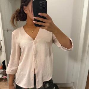 Ljusrosa blus från JDY med knappar - Superfin ljusrosa blus från JDY med v-ringning och knappar framtill. Skjortan har trekvartsärm och är gjord i ett lätt, krinklat tyg som känns luftigt och skönt. Perfekt för dig som gillar en enkel men stilren look.