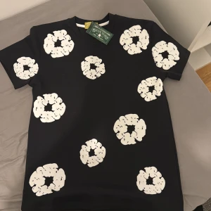 Denim tears tshirt - Cool svart t-shirt från denim tears med unika vita mönster tryckta över hela tröjan. T-shirten har klassisk passform och rund hals. Perfekt för dig som vill sticka ut med en stilren outfit. Den är helt ny och oanvänd.