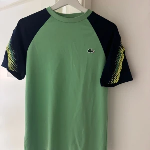 Grön Lacoste Sport t-shirt XS - Snygg grön t-shirt från Lacoste Sport med mörkblå ärmar och coolt mönster i gult och grönt på axlarna. Klassisk Lacoste-logga på bröstet. Regular fit och tillverkad i mjuk bomull, perfekt för en sportig look.