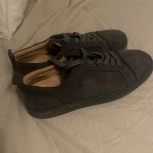 Mörkgrå sneakers från Christian Louboutin - Snygga mörkgrå sneakers från Christian Louboutin med klassisk rund tå och platt sula. Ovandelen är i mocka och skorna har snörning. Insidan är beige och yttersulan är röd med Louboutins signatur. Perfekt för dig som gillar exklusiva detaljer. Låda ingår.