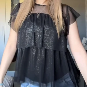 Svart glittrig blus med volanger - Säljer en svart glittrig blus med transparenta, korta volangärmar och lager på lager-effekt. Blusen har en luftig passform och glittrande detaljer som gör den extra snygg. 