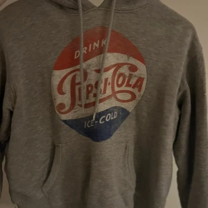Grå Pepsi hoodie med vintage-tryck - Grå hoodie från Pepsi med stort retro Pepsi-Cola tryck i rött, vitt och blått på bröstet. Tröjan har huva med snörning och en klassisk känguruficka framtill. Mjuk insida och ribbade muddar vid ärmslut och nederkant. Perfekt för dig som gillar ikoniska loggor.
