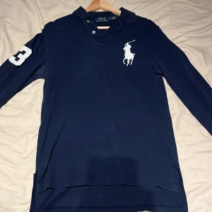 Marinblå långärmad pikétröja Ralph Lauren - Tja, Säljer en marinblå långärmad pikétröja från Polo Ralph Lauren, custom fit. Tröjan har klassisk krage, knappar vid halsen och stort vitt polospelar-logotyp på bröstet samt en vit siffra 3 på ärmen. Perfekt för dig som gillar sportig och stilren look.