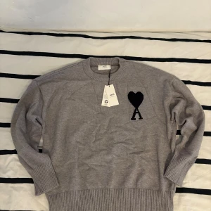 Grå stickad tröja från Ami - Snygg grå stickad tröja från Ami Alexandre Mattiussi med svart hjärta och A broderat på bröstet. Tröjan har rund hals, ribbade muddar och är långärmad. Perfekt för dig som gillar stilrena detaljer och vill ha något som sticker ut lite. Hör av dig om du har några frågor.