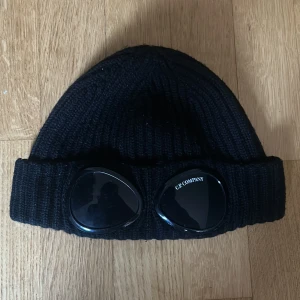Svart ribbad mössa från C.P. Company - Svart ribbstickad mössa från C.P. Company med ikonisk linsdetalj på uppviket. Snygg och stilren design som passar perfekt till streetwear. Materialet är stickad ull och mössan har en klassisk form med bred uppvikt kant. Lite små repor på glaset och jag har rivit bort en lapp inuti mössan som man ser på bild 3, annars är den i riktigt bra skick