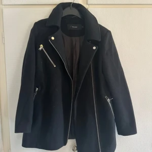 Svart ulljacka med dragkedjor Vero Moda - Stilren svart jacka från Vero Moda i ullblandning med coola silvriga dragkedjor och bred krage. Jackan har asymmetrisk dragkedja framtill och två snedställda fickor med dragkedja. Perfekt för dig som gillar en edgy look.