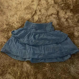 Blå volangkjol med resårmidja - Supersöt blå kortkjol med bred resår i midjan och dubbla volanger. Kjolen har en luftig och flowy känsla, perfekt för dig som gillar en avslappnad men ändå trendig stil. Materialet känns lätt och skönt mot huden.