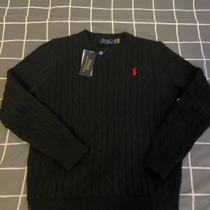 Svart stickad tröja Polo Ralph Lauren - Svart kabelstickad tröja från Polo Ralph Lauren i 100% bomull. Klassisk rund halsringning, långärmad modell och röd broderad logga på bröstet. Perfekt för dig som gillar stilrena och tidlösa plagg med en sportig touch.