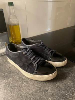 Svarta lanvins 41  - Snygga svarta sneakers med ovandel i mocka och en glansig, lackad tå. Skorna har vita sulor och klassisk snörning. Perfekta för dig som gillar en stilren men ändå unik look. Storlek 41 