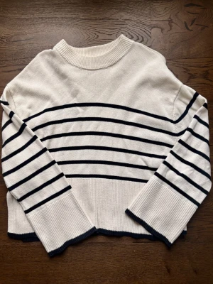 Vit stickad tröja med marinblå ränder - Snygg vit stickad tröja med marinblå horisontella ränder och bred ribbad mudd vid ärmslut och hals. Tröjan har en avslappnad passform och klassisk rund halsringning. Perfekt för en stilren och tidlös look.