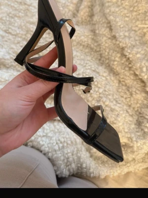 Svarta lackpumps med remmar - Snygga svarta pumps i lack med smala remmar och fyrkantig tå. Skorna har en hög klack med modern form och öppen häl. Perfekta för att lyfta din outfit med en trendig touch.