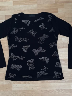 Svart långärmad topp med fjärilar - Snygg svart långärmad topp från med silverglittriga fjärilar över hela framsidan. Toppen har en klassisk rund halsringning och är tillverkad i ett mjukt, stretchigt material som sitter bekvämt på kroppen. Bröst 100cm, används en gång.