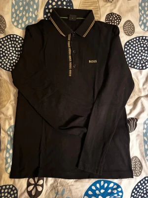 Svart långärmad piké från BOSS - Svart långärmad pikétröja från BOSS med gulddetaljer på kragen och knappraden. Klassisk krage med smala ränder och BOSS-logga broderad på bröstet. Skön och stilren modell som passar perfekt till jeans eller chinos.