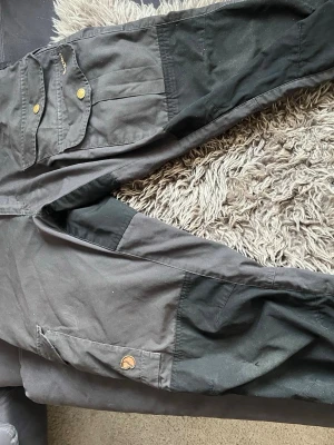 Svarta cargobyxor från Fjällräven - Snygga svarta cargobyxor från Fjällräven med förstärkta partier på knän och bak, stora fickor med tryckknappar och broderad G-1000-logga. Byxorna har klassisk cargomodell och är tillverkade i slitstarkt material, perfekta för äventyr och outdoor. OBS liten reva på höger knä