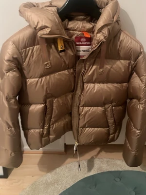 Beige dunjacka från Parajumpers - Snygg beige dunjacka från Parajumpers med kort modell, stor huva och dragkedja framtill. Jackan har två stora fickor och coola detaljer som gul rem vid halsen. Perfekt för kalla vinterdagar och riktigt trendig look.