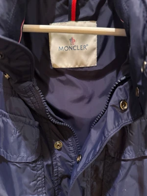 Moncler fieldjacket - Snygg mörkblå vindjacka från Moncler med klassisk logga på ärmen. Jackan har flera fickor med dragkedja, axelklaffar och hög krage. Tillverkad i polyester med detaljer i metall och ribbade muddar. Perfekt för dig som vill ha en stilren och sportig look.