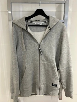 Grå zip-hoodie från Björn Borg - Snygg grå hoodie från Björn Borg med dragkedja framtill, huva med dragsko och två stora fickor. Mjuk bomullsblandning och diskret logga på ena fickan. Perfekt för en avslappnad och sportig stil. Aldrig använd dm för frågor!