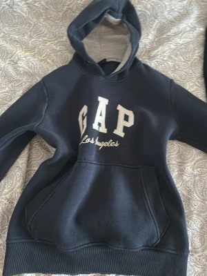 Mörkblå GAP hoodie Los Angeles S - Snygg mörkblå hoodie från GAP med stor vit logga och texten 'Los Angeles' på bröstet. Hoodien har en stor känguruficka framtill och ribbade muddar. Insidan av huvan är ljusgrå och mjuk. Perfekt för chill dagar och streetwear vibes.