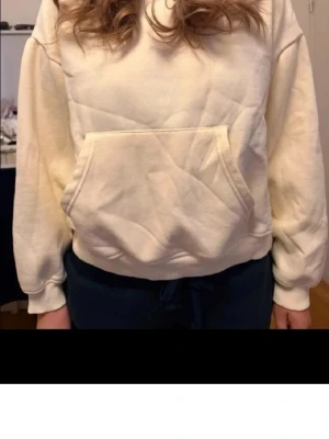 Krämvit hoodie med magficka - En enkel och clean ljusgul hoodie med stor magficka och huva. Hoodien har ribbade muddar vid ärmslut och nederkant, samt en avslappnad passform. Perfekt för chill dagar eller när du vill ha något bekvämt och stilrent. Materialet känns mjukt och skönt mot huden.