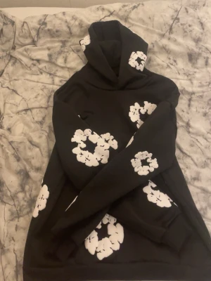 Denim tears hoodie - Säljer en svart hoodie med stora vita blommönster tryckta över hela tröjan, inklusive huvan och ärmarna. Hoodien har en klassisk passform med ribbade muddar och en stor huva. Perfekt för dig som vill sticka ut med en unik och trendig stil.