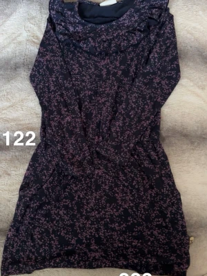 Mörkblå blommig långärmad klänning - Mysig mörkblå klänning med lila blommönster och långa ärmar. Klänningen har, avslappnad passform och en hög krage. Perfekt för dig som gillar bekväma och snygga plagg med fina detaljer.🌸 Fint använt skick. Storlek 122 