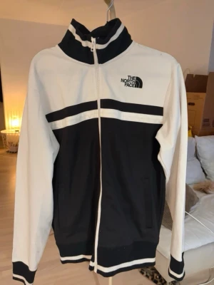 Svartvit ziptröja från The North Face - Snygg svartvit ziptröja från The North Face med hög krage och dragkedja hela vägen. Tröjan har vita ärmar, svarta partier framtill och baktill samt vita och svarta ränder över bröstet och vid muddar. Perfekt för en sportig och stilren look.