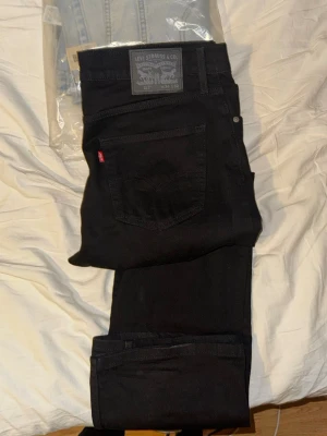 Svarta Levi's jeans med rak passform - Säljer ett par svarta jeans från Levi's med klassisk rak passform och fem fickor. Jeansen har den ikoniska röda Levi's-lappen på bakfickan och är tillverkade i ett slitstarkt bomullsmaterial. Perfekta för en clean och stilren look.