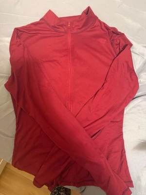 Röd sporttröja med dragkedja - Säljer en röd långärmad sporttröja med halv dragkedja framtill. Tröjan har en hög krage och är tillverkad i ett stretchigt funktionsmaterial som passar perfekt till träning eller löpning.