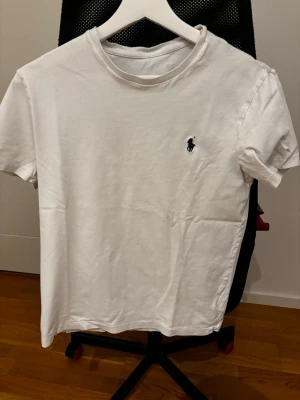 Vit t-shirt från Ralph Lauren - Klassisk vit t-shirt från Ralph Lauren med den ikoniska mörkblå polospelaren broderad på bröstet. T-shirten har rund halsringning och korta ärmar. Tillverkad i mjuk bomull för en skön känsla och enkel stil.