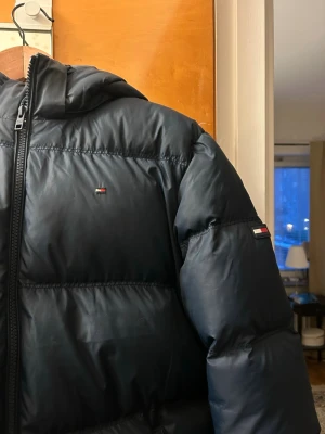 Svart dunjacka från Tommy Hilfiger - Snygg svart dunjacka från Tommy Hilfiger med huva och dragkedja. Jackan har diskreta loggor på bröstet och ärmen samt är fylld med dun och fjäder för extra värme. Perfekt för kalla vinterdagar och har en klassisk, stilren look.