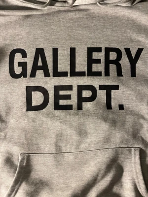 Grå hoodie från Gallery Dept. - Snygg grå hoodie från Gallery Dept. med stor svart text på bröstet. Klassisk känguruficka framtill, ribbade muddar och justerbar huva med dragsko. Tillverkad i mjukt bomullsmaterial som är perfekt för chill dagar. Använt den ett tag nu så därav priset