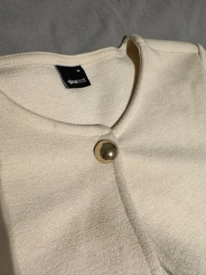 Krämvit kofta från Gina Tricot - Stilren krämvit kofta från Gina Tricot med rund halsringning och en stor, glansig guldfärgad knapp framtill. Koftan har en mjuk struktur och minimalistisk design som passar perfekt till många olika looks. Har aldrig använt då jag fick den som present men passade tyvärr inte.