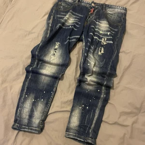 Mörkblå distressed jeans från DSquared2 - Säljer ett par mörkblå jeans från DSquared2 med slitningar, blekta partier och färgstänk för en riktigt edgy look. Jeansen har klassisk femficksmodell, låg midja och knappgylf. Perfekta för dig som gillar streetwear och vill sticka ut. Bra skick, pris går att diskutera