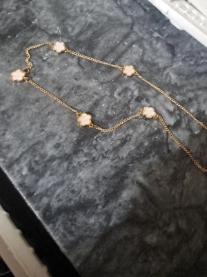 Guldfärgat halsband med blommor - Snyggt halsband med tunn guldfärgad kedja och fem vita blommor i fyrklöverform. Blommorna har en pärlemorskimrande yta och är jämnt placerade längs kedjan. Perfekt för dig som vill ha en elegant och trendig accessoar.