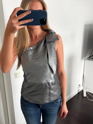 Glittrig silver topp med rosett - Snygg ärmlös topp i glittrigt silvermaterial med en stor rosett på ena axeln. Toppen har asymmetrisk axel och sitter figurnära. Perfekt för dig som vill sticka ut med en trendig look.