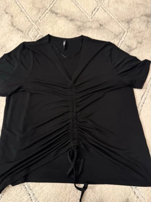 Svart topp med rynk och knytband - Trendig svart topp med korta ärmar och V-ringning. Framtill har toppen en snygg rynkning med knytband som ger en cool och justerbar look. Tillverkad i mjukt, stretchigt material som känns skönt. 